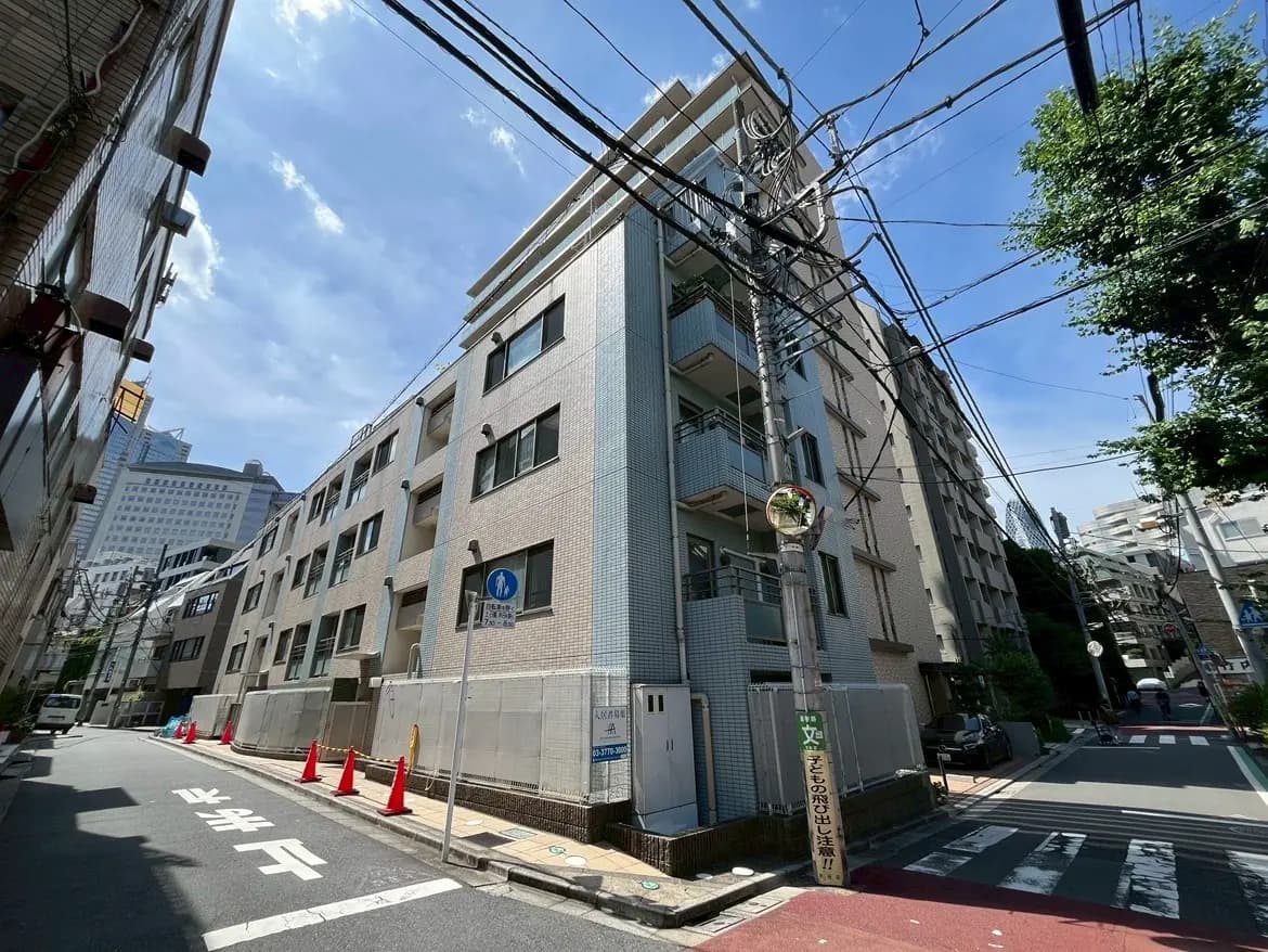 - 都市の住宅街に位置するアパートメントビルの外観、青空と電柱が特徴的な風景、周囲には赤いコーンと通行人の姿が見える