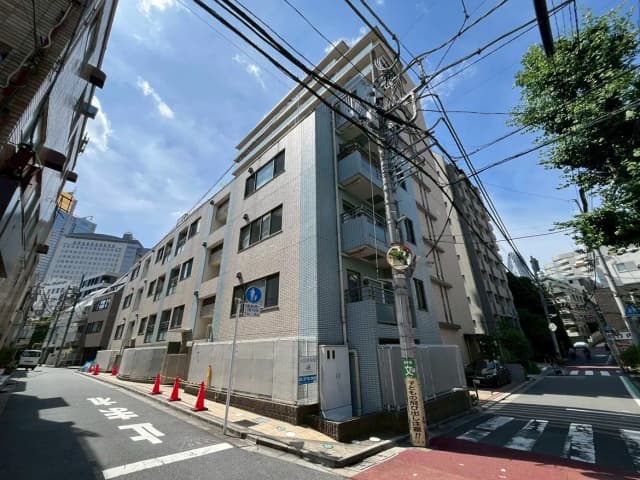 - 都市の住宅街に位置するアパートメントビルの外観、青空と電柱が特徴的な風景、周囲には赤いコーンと通行人の姿が見える