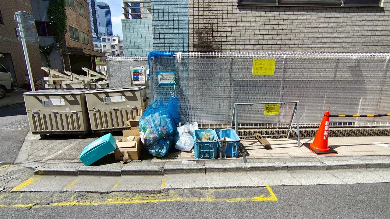 - 街の景観とごみ置き場、ペットボトルや紙ごみの入った袋、プラスチック容器が並ぶ街道沿いの風景