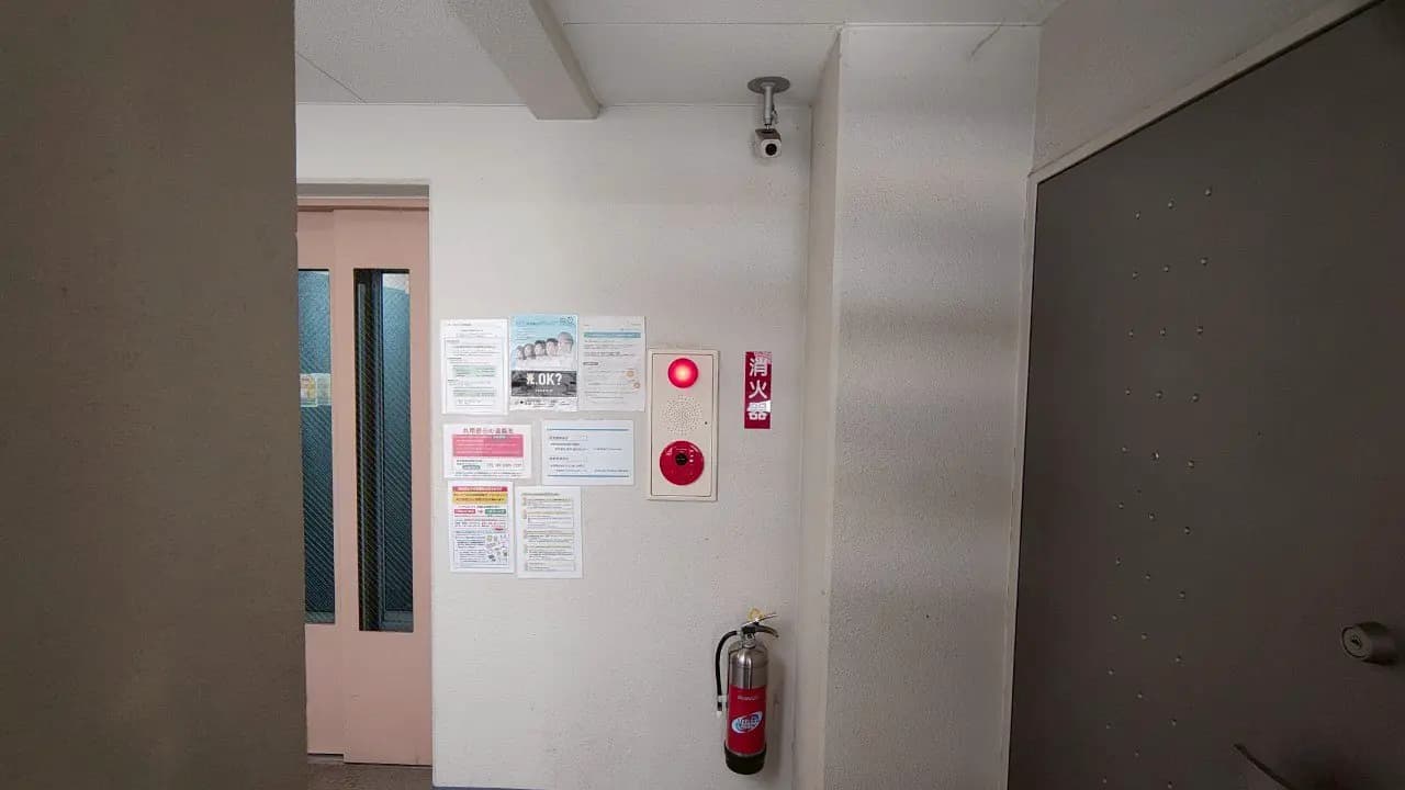 - 廊下の壁に貼られた各種掲示物と赤い非常ベル、消火器が設置された防火基本設備の一角