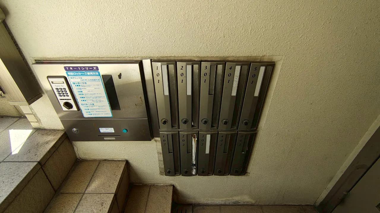 - アパートの入口に設置されたインターホンと複数の郵便受け、番号が刻まれたスタイリッシュな金属製のデザイン