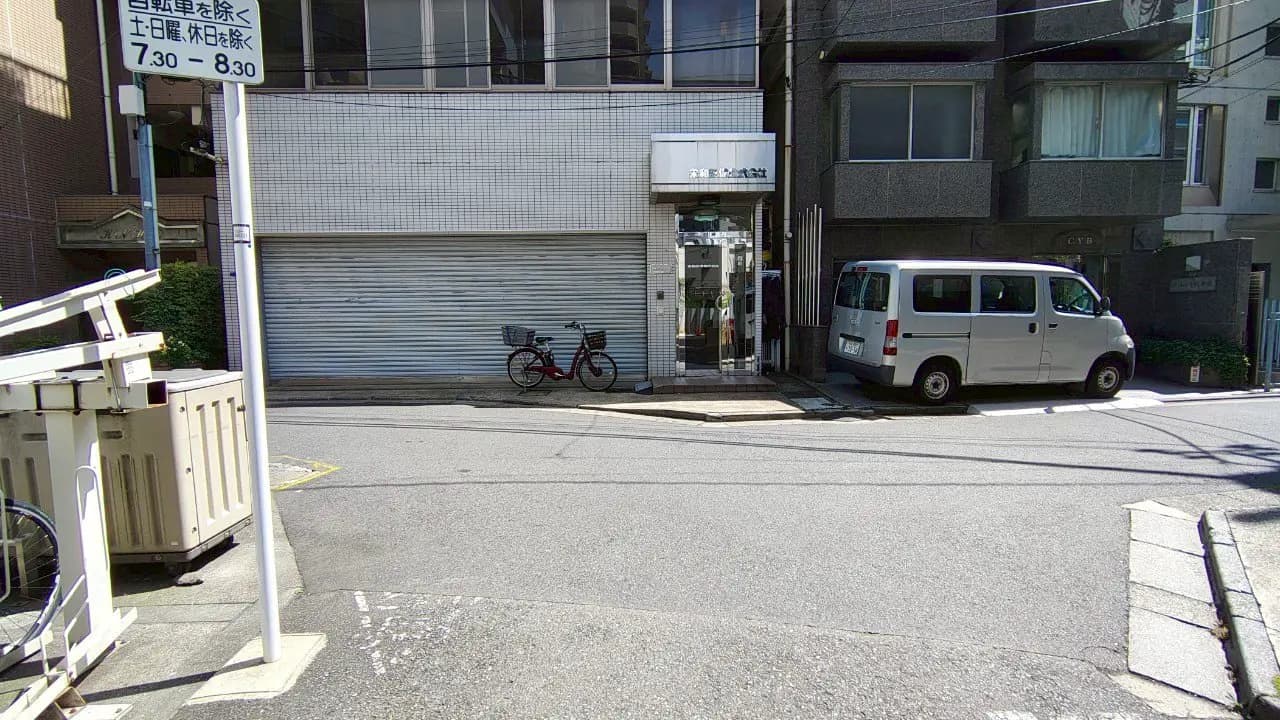 - 近隣の建物と白いバン、赤い自転車が置かれた通りの風景、シャッター付きの商業施設の入口