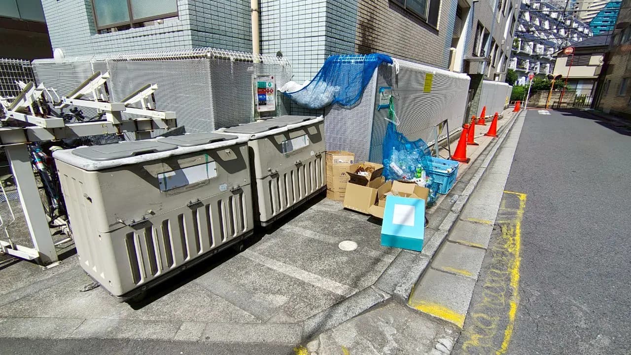 - 都市の裏道に置かれたゴミ箱、周囲には段ボールやプラスチックゴミが散乱し、作業中の設備が見える風景