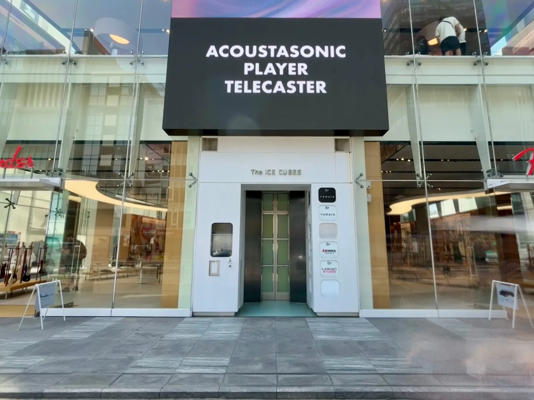 - 斬新なデザインのビルの入り口、看板には「ACOUSTASONIC PLAYER TELECASTER」の文字、ガラス張りの外観と現代的なエレベーターのエントランス