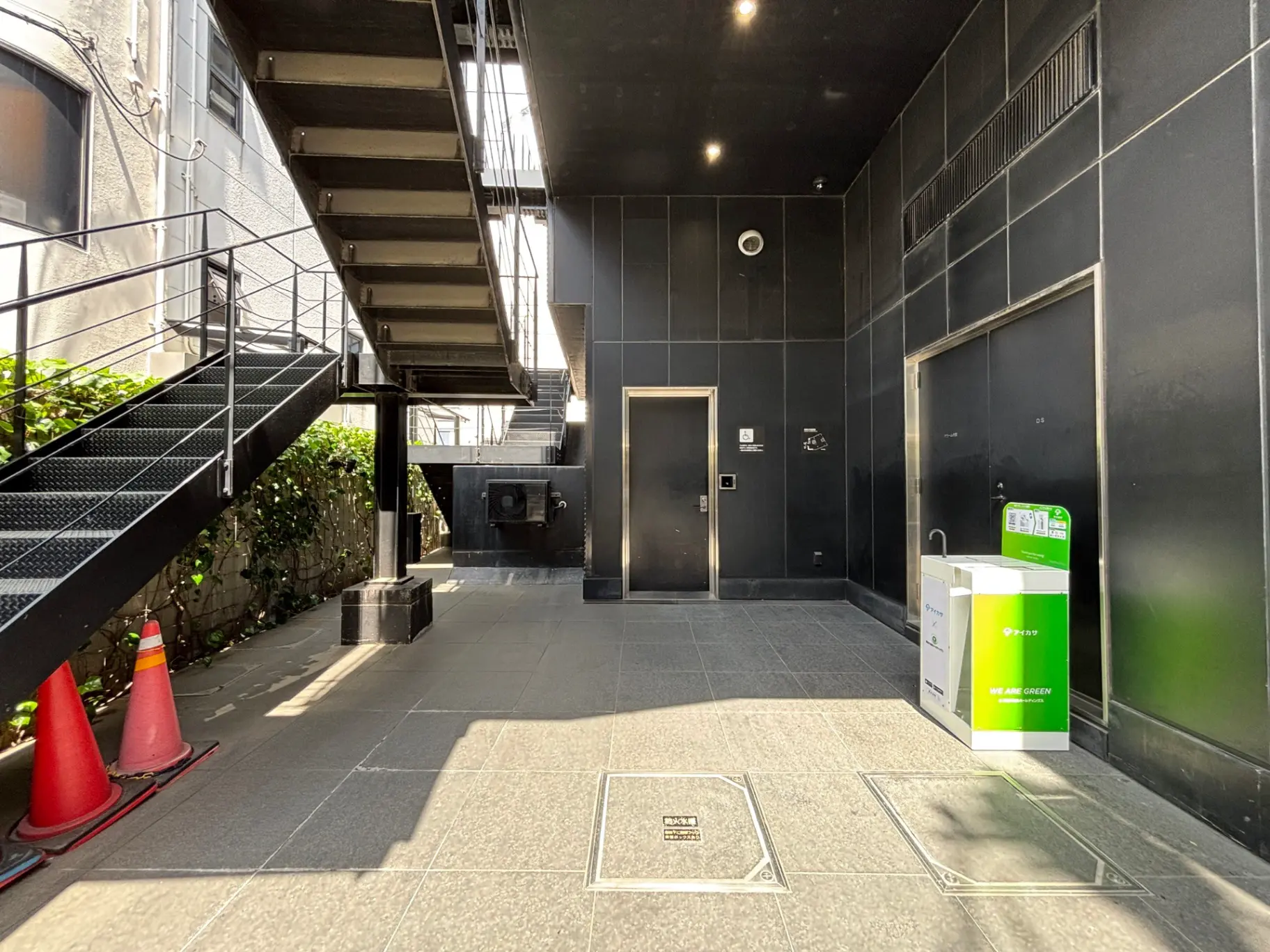 - 黒い外壁の建物前にある金属製の階段、周囲に配置されたコーンとゴミ箱、日差しが差し込む現代的なエントランス