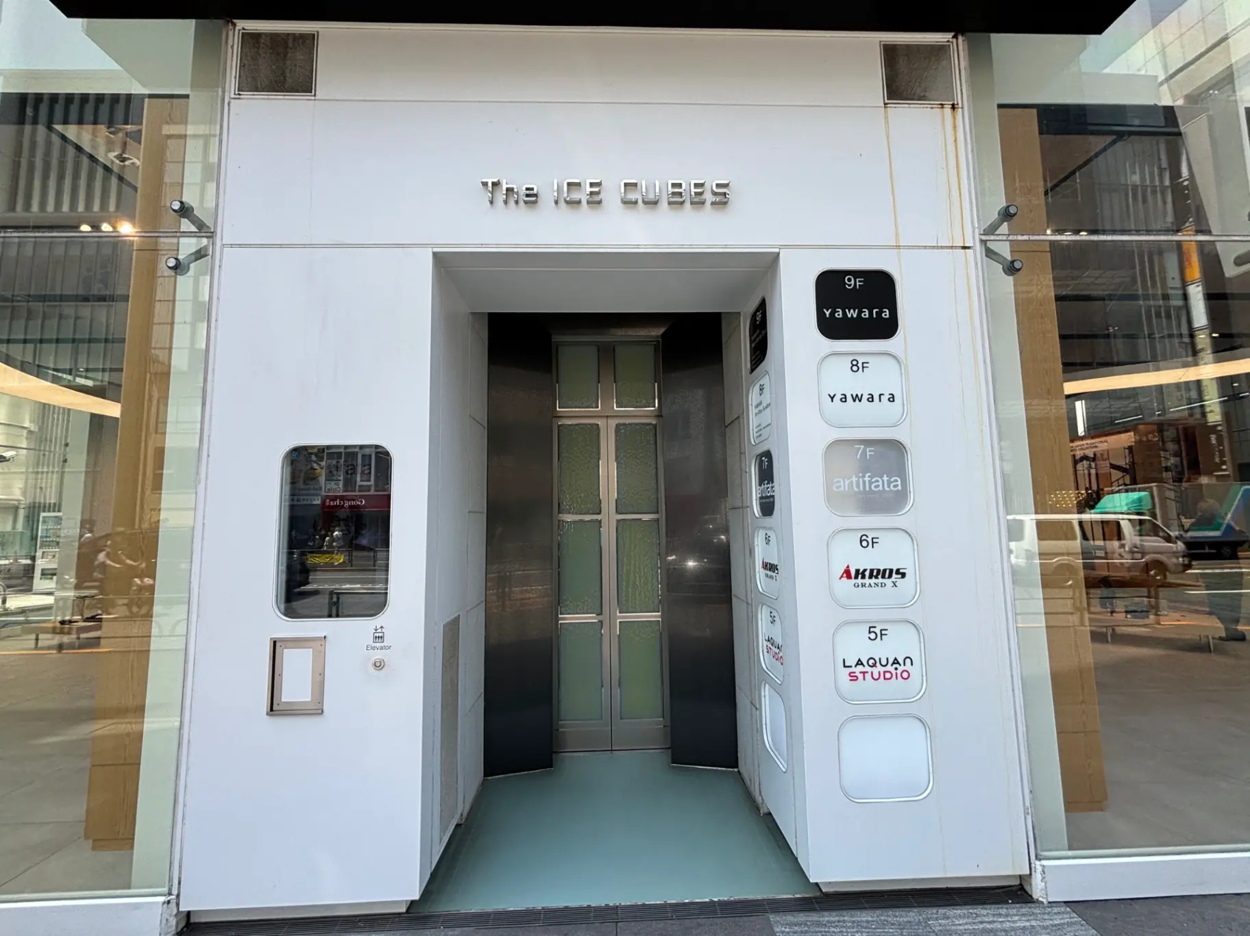 - モダンなビルのエレベーター入口、ホワイトの外壁に「The ICE CUBES」の文字、各階の店舗名を表示した案内板が特徴的なデザイン