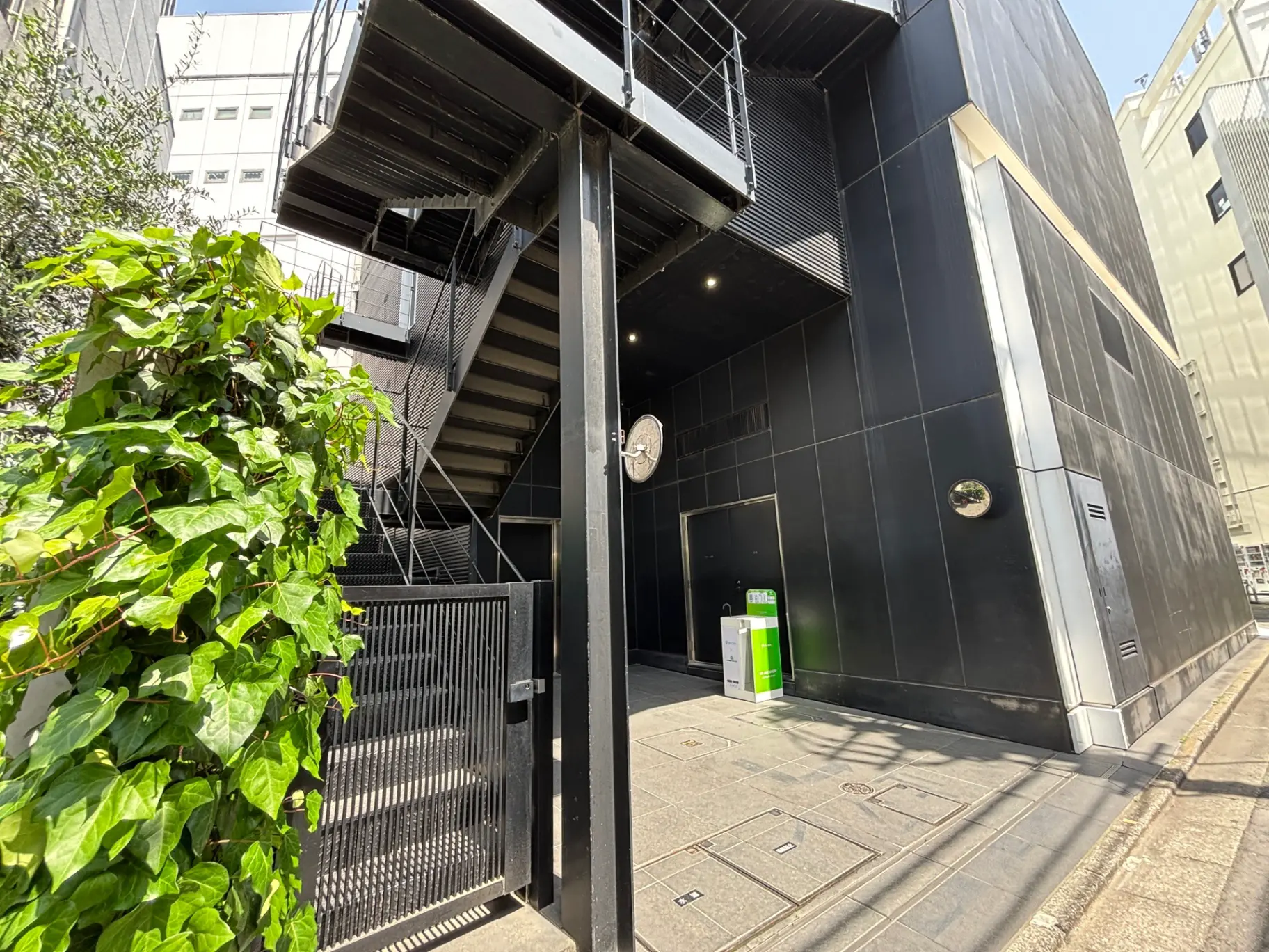 - 現代的なデザインの黒い外壁のビルと鉄製の階段、並ぶ緑の植物、道沿いの景観