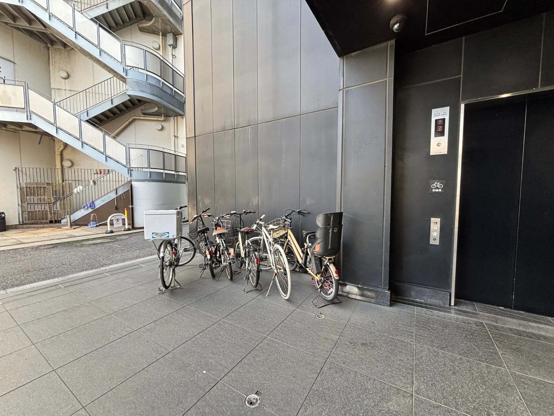 - 建物の入口前に並んでいる自転車、周囲には階段とエレベーターがあるモダンな都市景観