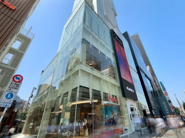 - 透明なガラス外壁を持つ現代的な商業ビル、青空の下で賑わう通りの様子と店舗の看板が映える景観