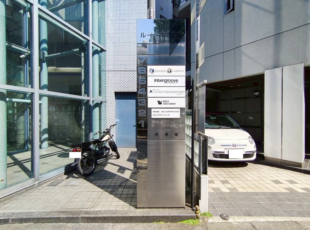 - 近代的なビルの入り口看板、企業名が表示された金属製の標識と横に停車したバイクと自動車