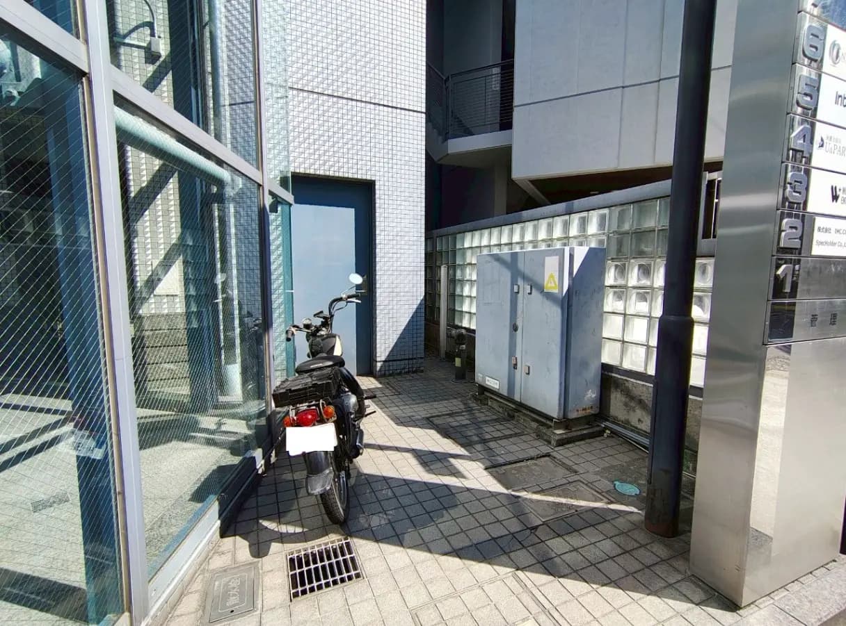 - ビルの横に停まっているバイク、ガラス張りの壁と青いドアのある通路の風景、隣には電気設備のボックスがある
