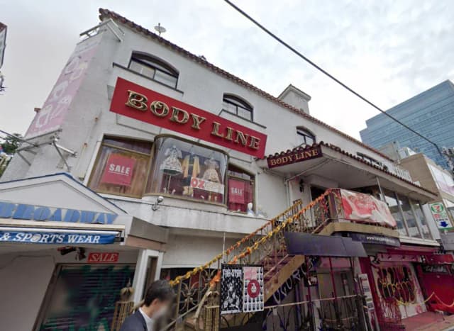 - BODY LINEの店舗外観、セールのポスターが貼られたガラスショーウィンドウ、階段と周辺の商業施設が見える風景