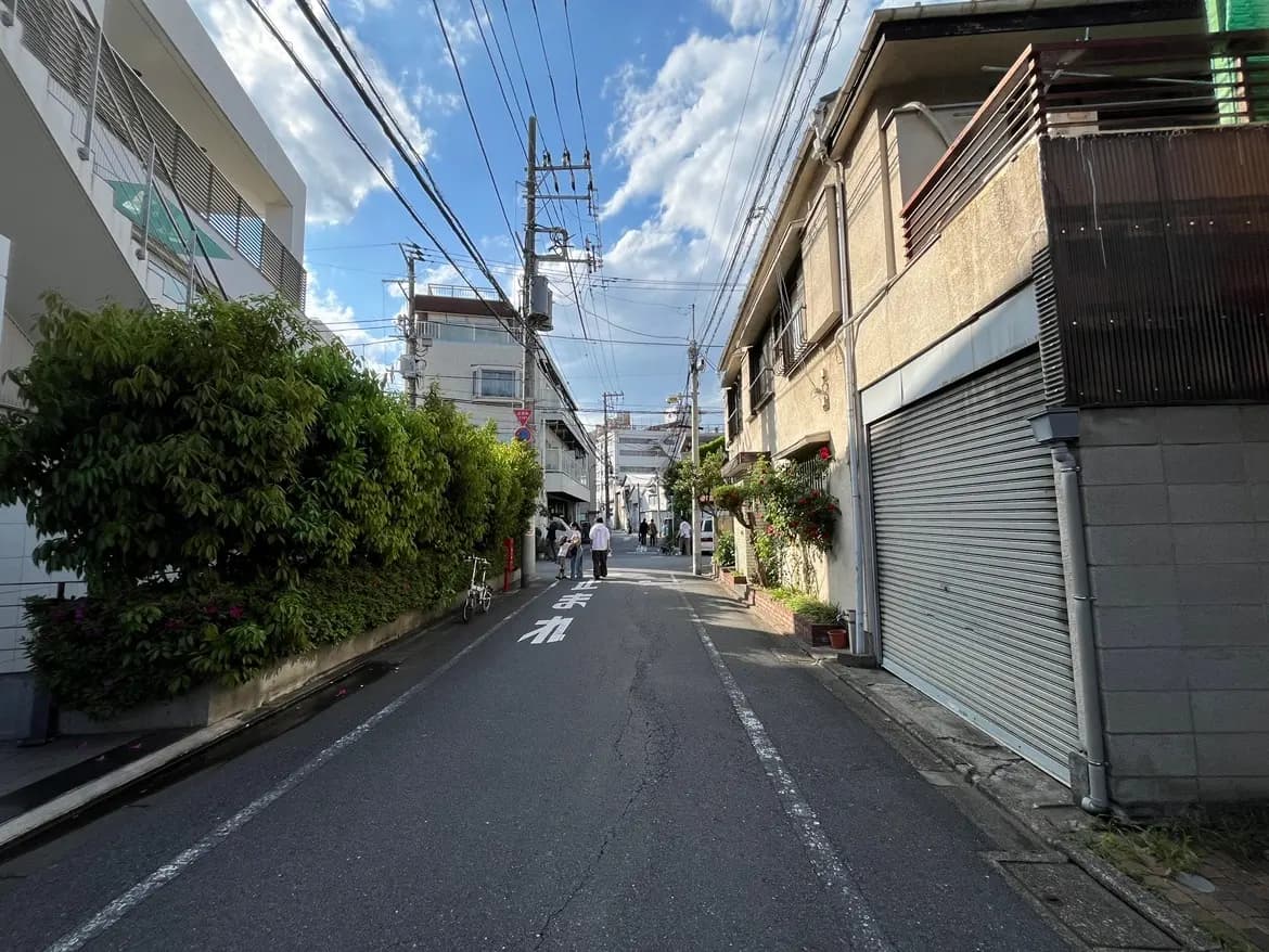 - 都会の静かな路地、両側には低い建物と緑が茂った植え込み、青空に続く道路の景観