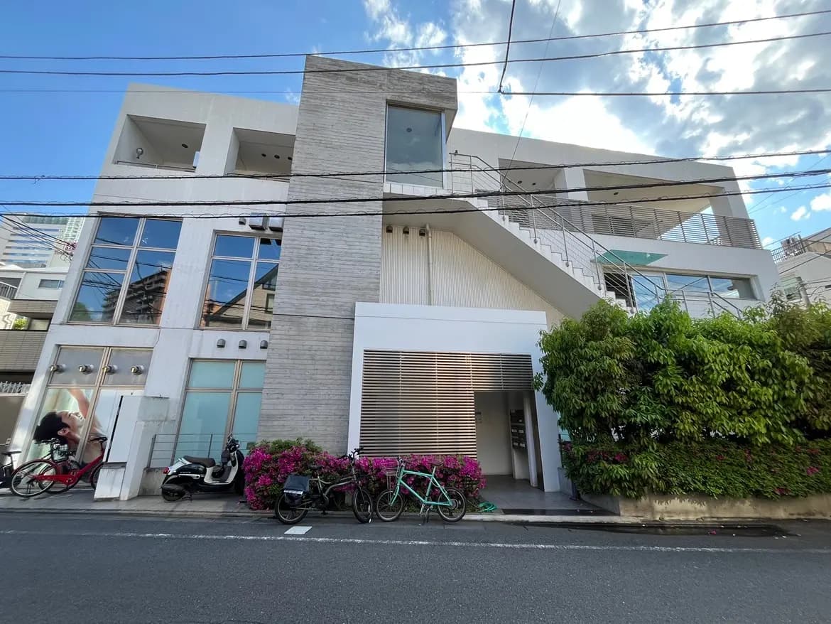 - モダンな外観のビル、ガラス張りの窓と階段、周囲にバラ色の花が咲く緑豊かな植栽、道に停まる自転車とバイク