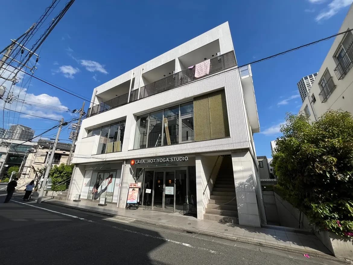 - LAVAホットヨガスタジオのモダンな外観、青空を背景にした建物と周囲の街並み、看板と駐車スペースが見える街角の風景