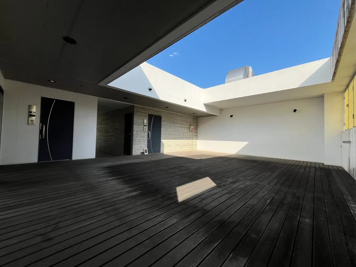 - 明るい空の下、シンプルでモダンなデザインの空間、木製デッキとコンクリート壁が特徴のエントランスエリア