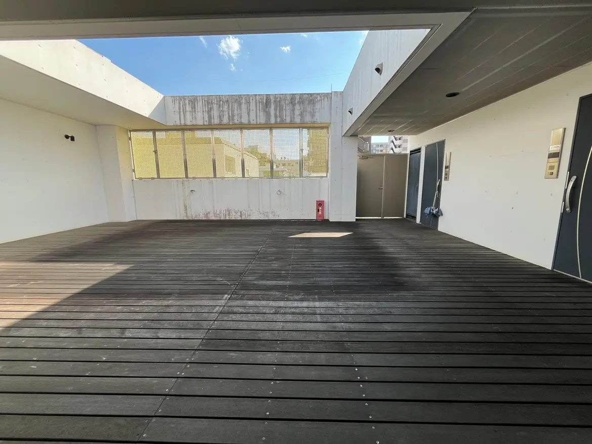 - 明るい青空の下に広がる屋上テラス、木製のデッキ床と白い壁、周囲にはエントランスと窓のある現代的なデザインの空間
