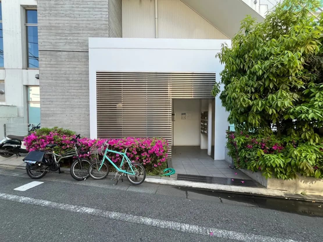 - 鮮やかな花に囲まれた現代的な建物の入口、左側に自転車とバイクが並ぶ風景