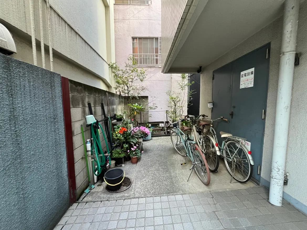 - 自転車が置かれた住宅の裏側、色とりどりの花が植えられたプランターと庭道具が並ぶエリア