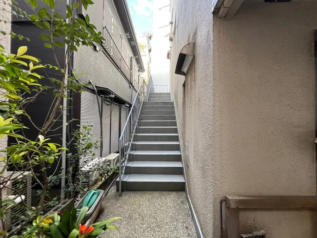 - 錆びた手すりの金属階段がある住宅の横道、緑豊かな植物と細長いアプローチの風景