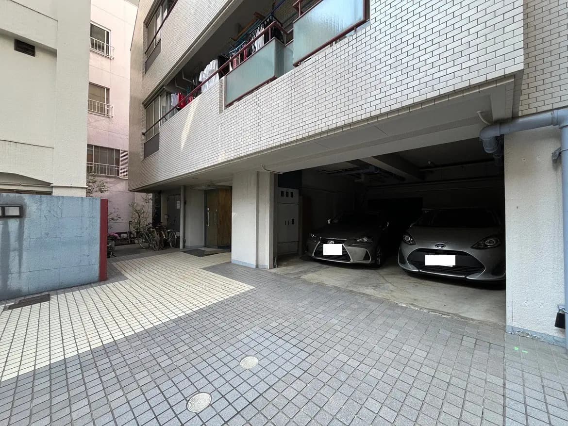 - 駐車場に停まっているシルバーの車2台と、その周辺のタイル舗装されたエリア、左右に建物が隣接する風景