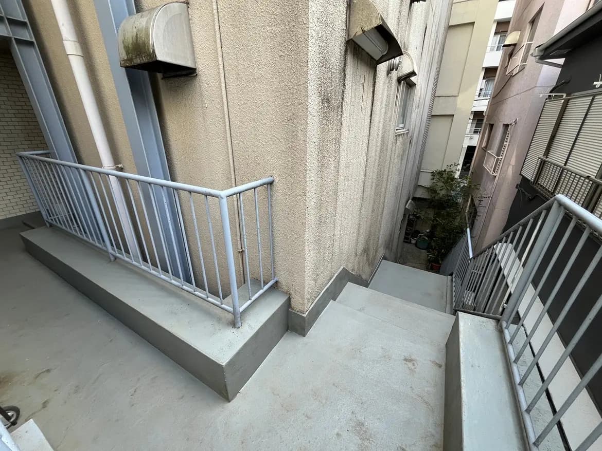 - 狭い通路の脇にある手すり付きの階段、周囲には高い建物が立ち並び、空間の奥に緑が見える風景