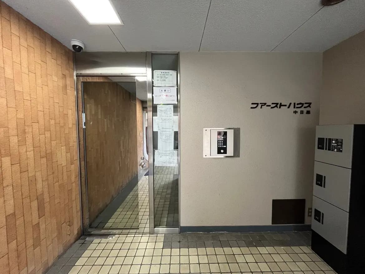 - 入口ドアとインターホン、壁に掲示されたファーストハウスのサイン、タイル張りの floor ツートンカラーの仕上げのエントランス