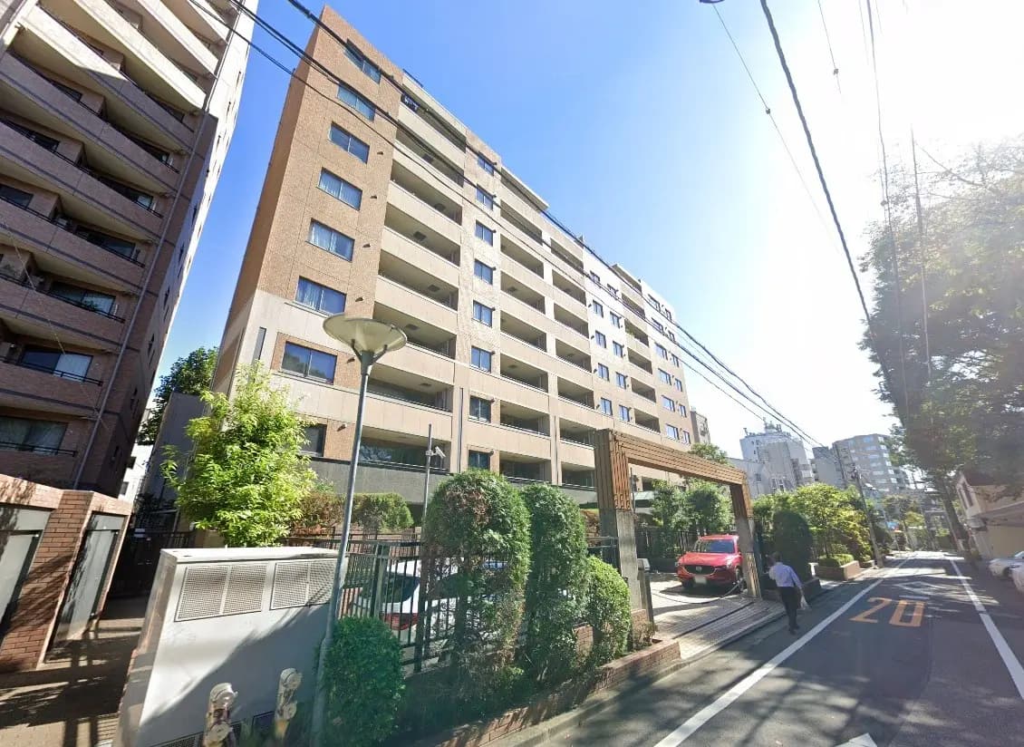 - 街中にある現代的なマンションの外観、周囲に緑の植栽と駐車場が見える晴れた日の風景