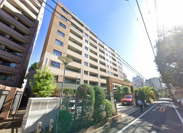- 街中にある現代的なマンションの外観、周囲に緑の植栽と駐車場が見える晴れた日の風景