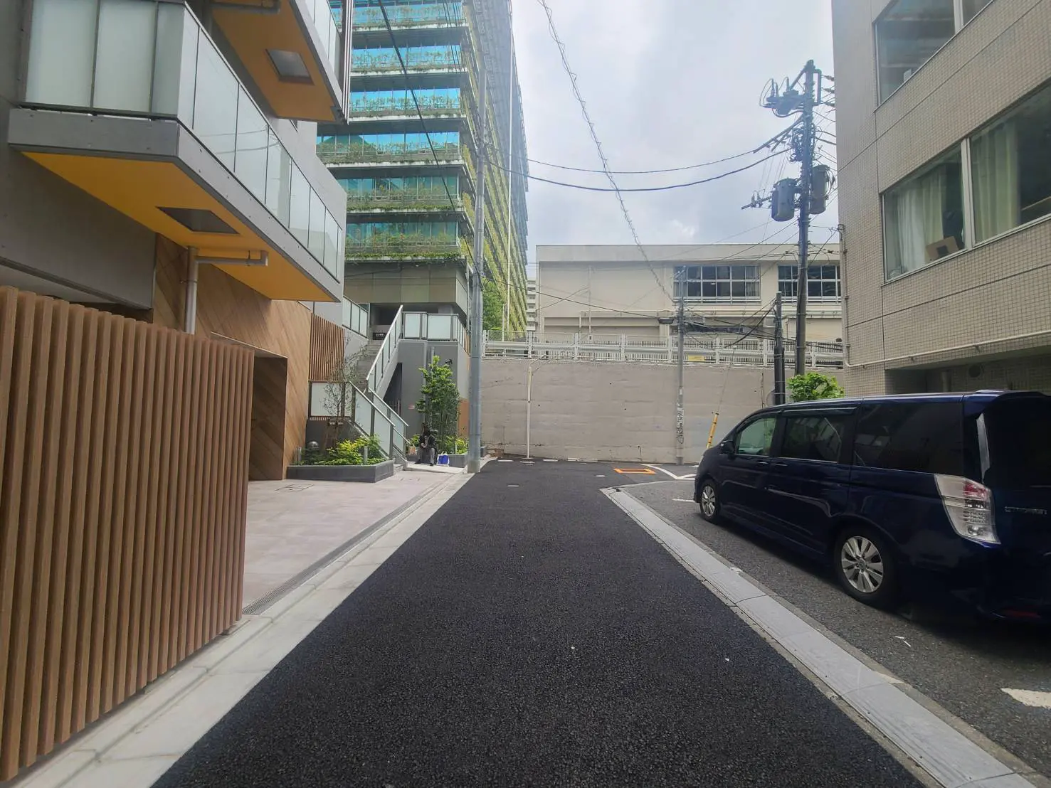 - モダンな都市の街路、左側に木材のフェンスと緑の植栽、右側に黒いミニバンが停車している景観