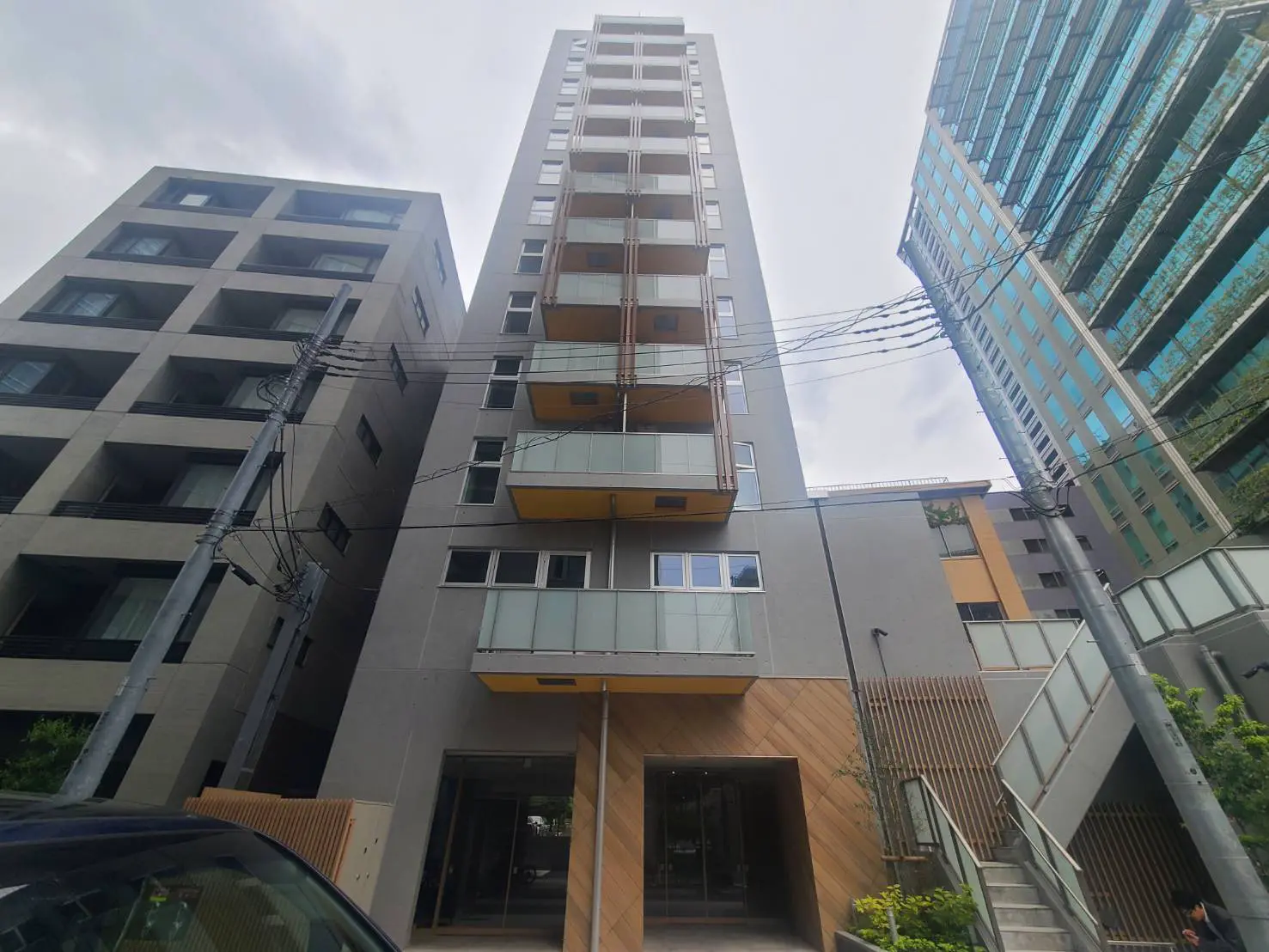 新しい高層マンションの外観、ガラスバルコニーと木目調のアクセントが特徴、周囲には他の現代的なビルが立ち並ぶ景観