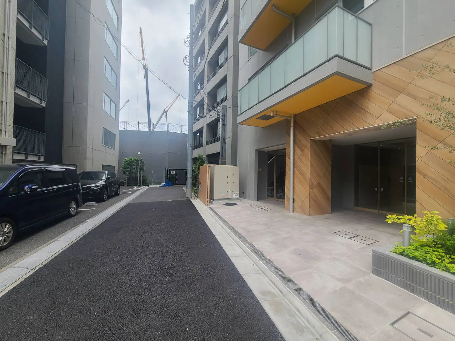- 現代的なマンションのエントランスと周辺の道路、左側には黒い車が並び、右側には木目調のデザインの壁とガラスのバルコニーが見える風景
