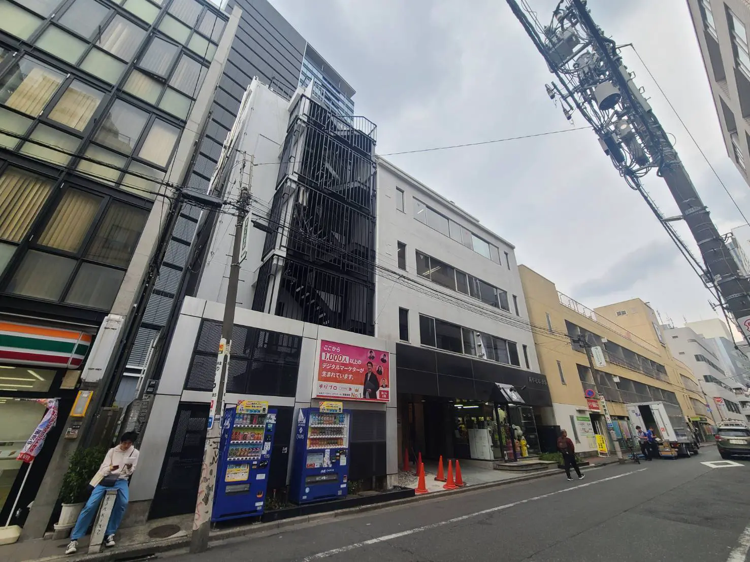 都市のビル群と歩道、ビルのファサードにチラシが掲示され、飲料自販機が並ぶ街の風景