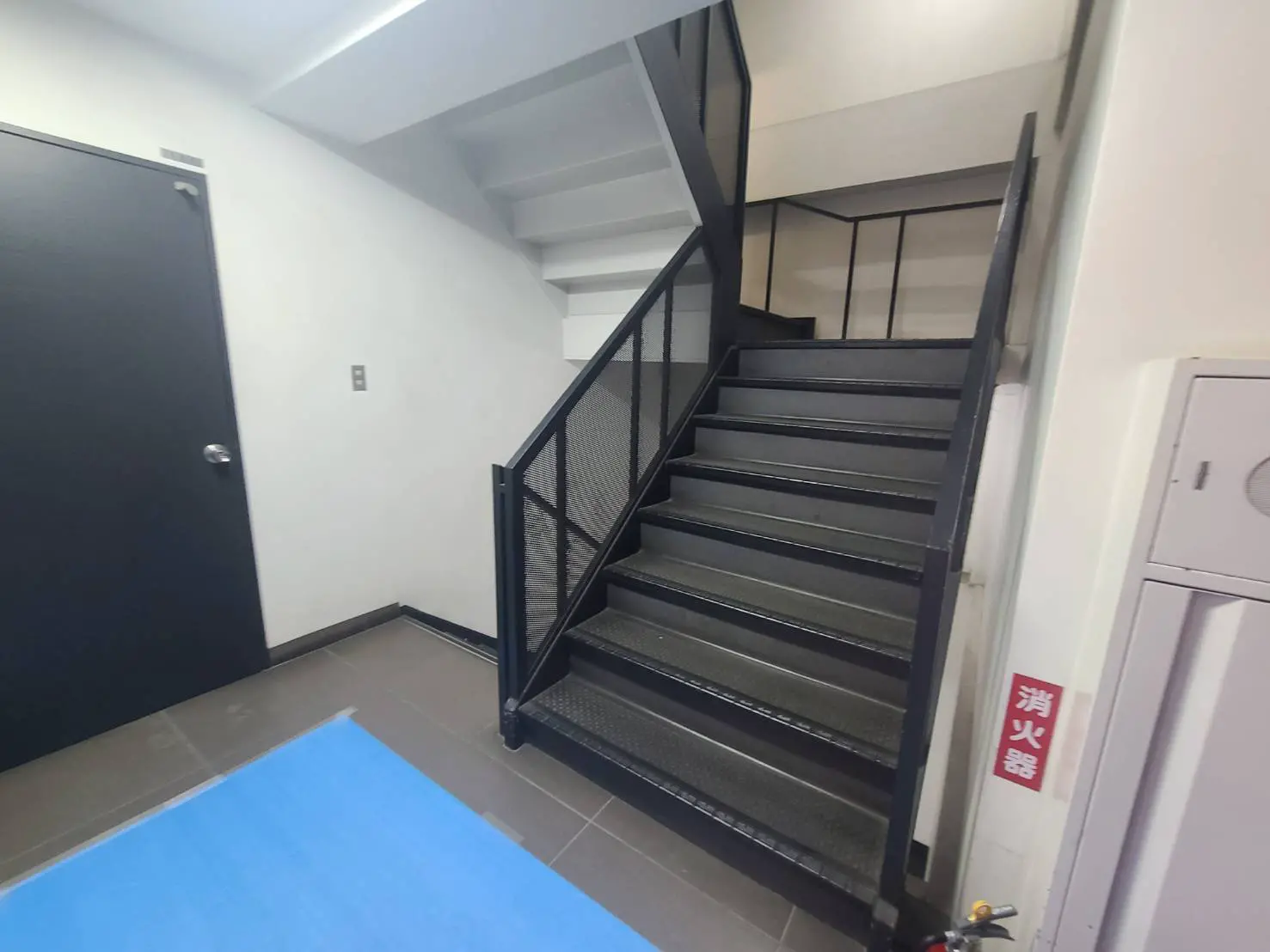 - モダンな階段と黒いドア、シンプルな内装のエントランススペース、青いマットと消火器が配置された写真