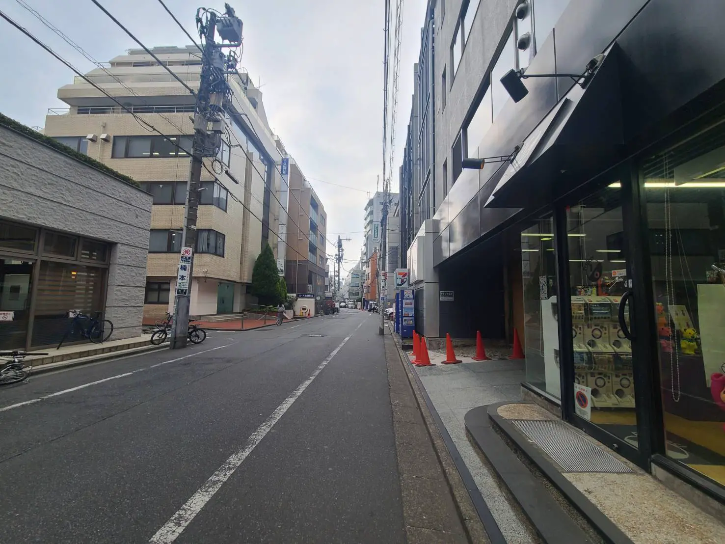 - 都市のビル群と静かな道路、手前に黄色のコーンが並ぶ風景、対向車線側には自転車が停められた様子
