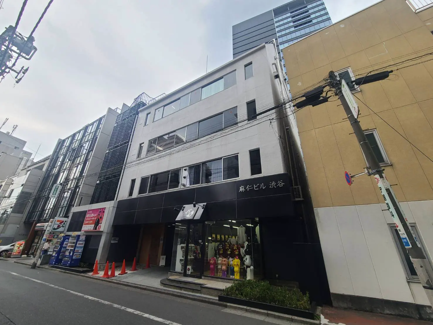 - 渋谷にある麻仁ビルの外観、近代的なデザインと黒い外壁、周囲に立ち並ぶビルと交通標識が見える通りの風景