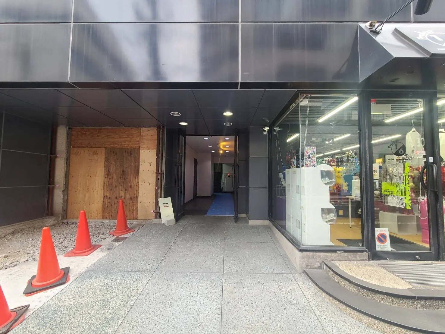 - 黒い外壁の商業ビルのエントランス、ガラスのショーウィンドウ、明るい照明が店内を照らす、土木工事中を示す簡易ボードとオレンジのコーンが配置された通路