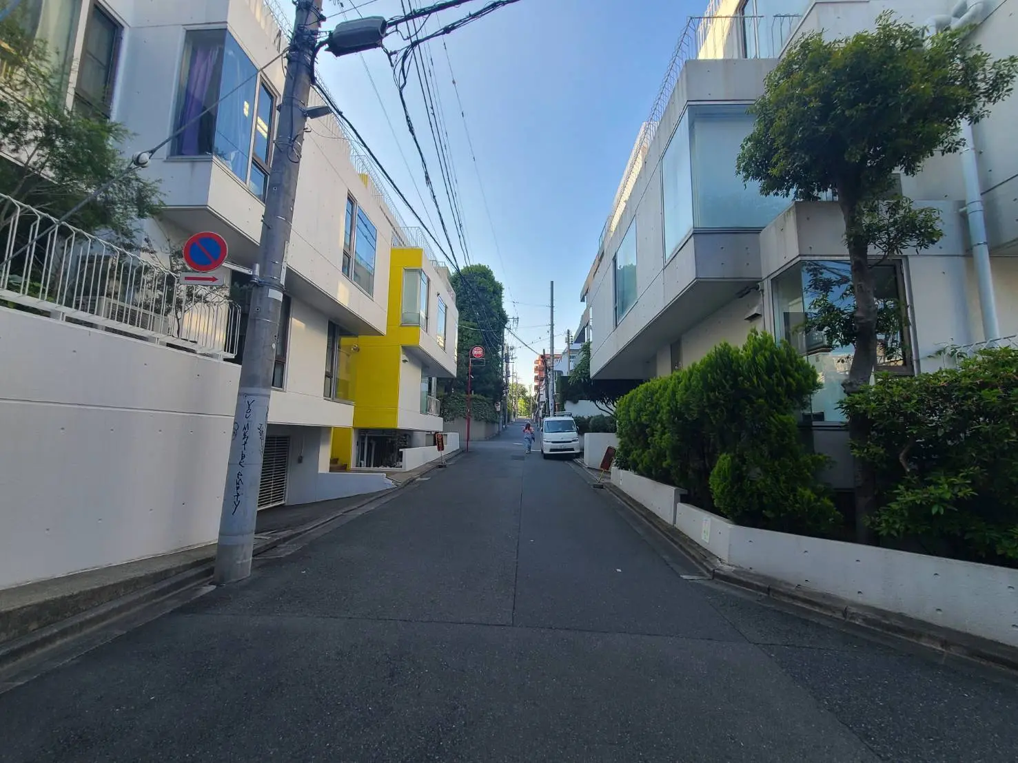 - 現代的な住宅街の風景、白い建物と明るい黄色のアクセント、緑の樹木と狭い道路が続く情景