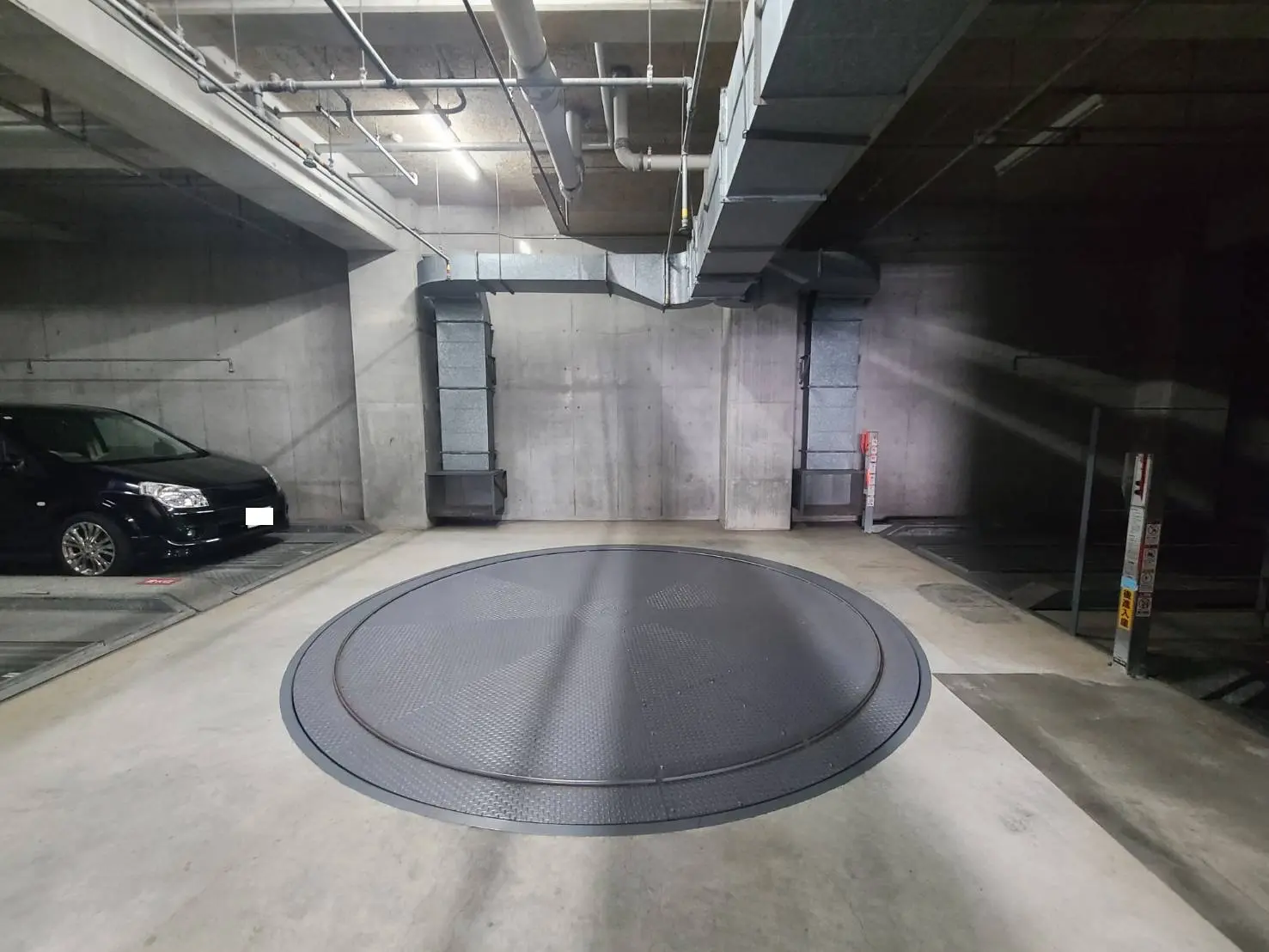 - コンクリート製の駐車場内に設置された車両回転プラットフォーム、左側に黒い車が停車している画像