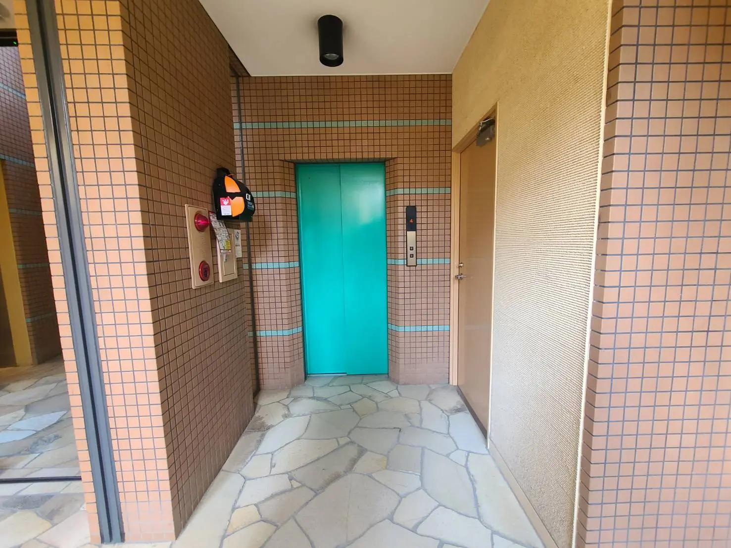 - モダンなエレベーターがある建物のエントランスホール、ブラウンのタイルの壁と石畳の床デザイン、インターホンや防災ベルが取り付けられたエリア