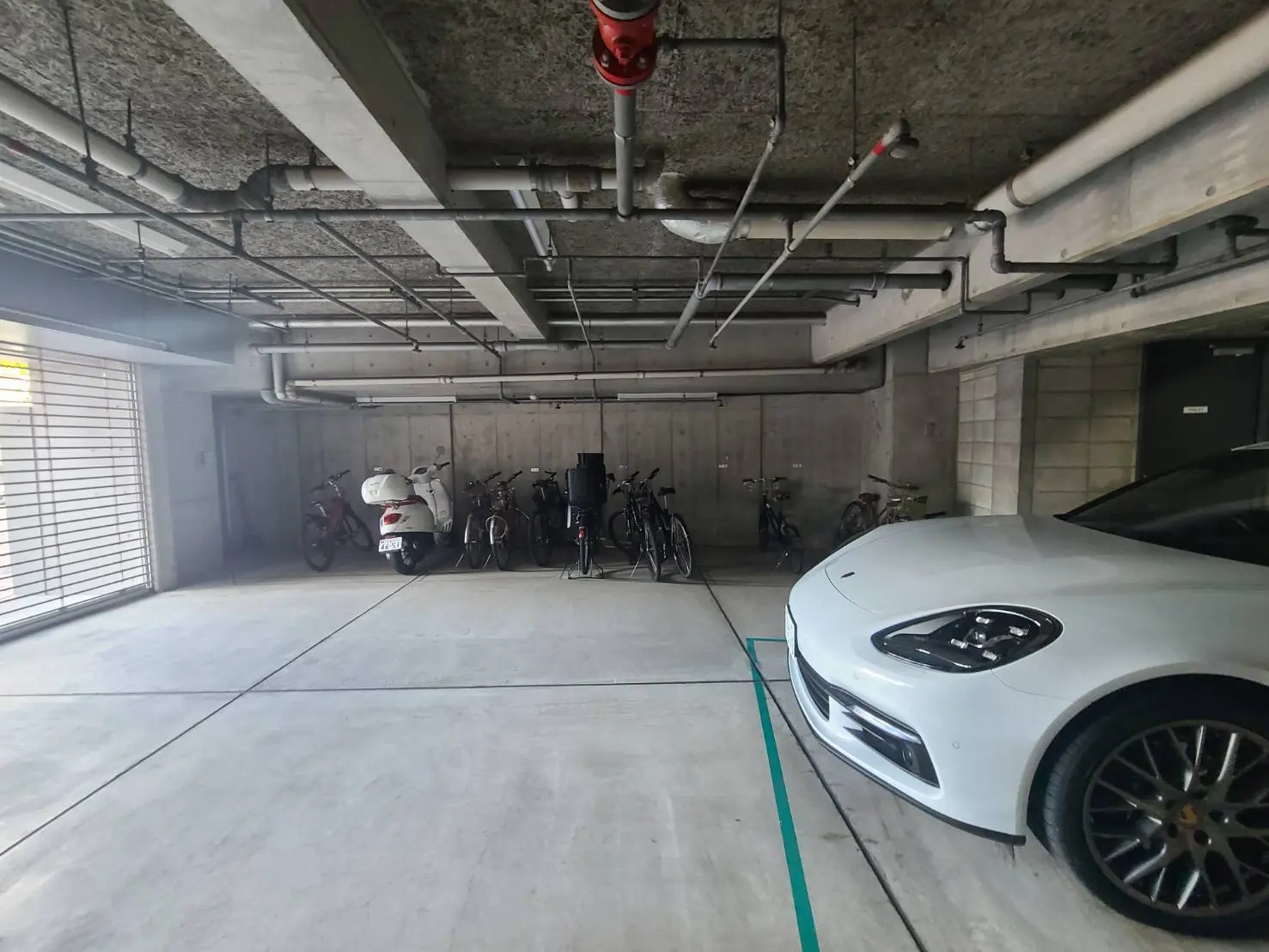 - コンクリート製の駐車場に停められた白いスポーツカーと様々な自転車が並ぶ様子、壁には配管が見える工業的なインテリア