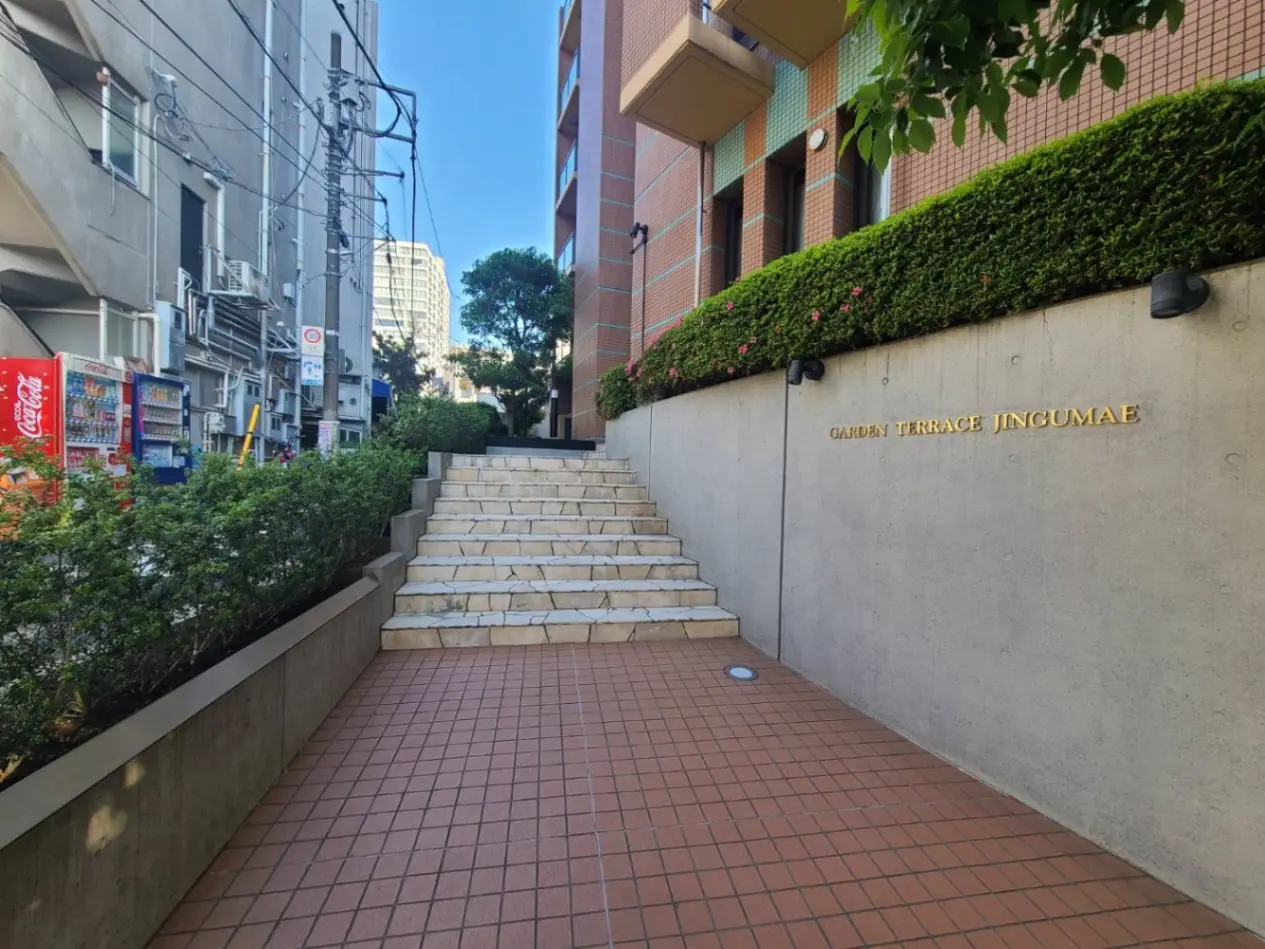 - 街の風景にある「GARDEN TERRACE JINGUMAE」の看板と整備された階段、周囲の緑と近くの自販機があるエントランスの様子