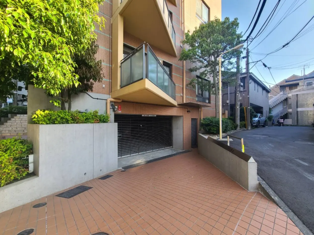 - 都市の静かな通りに面したモダンなマンションのエントランス、周囲には緑の植栽とタイル敷きの歩道が広がっている風景