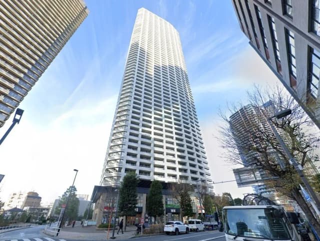 - 高層ビルの外観、周囲に並ぶ近代的な建物と青空、街路樹や車両が見える都市風景