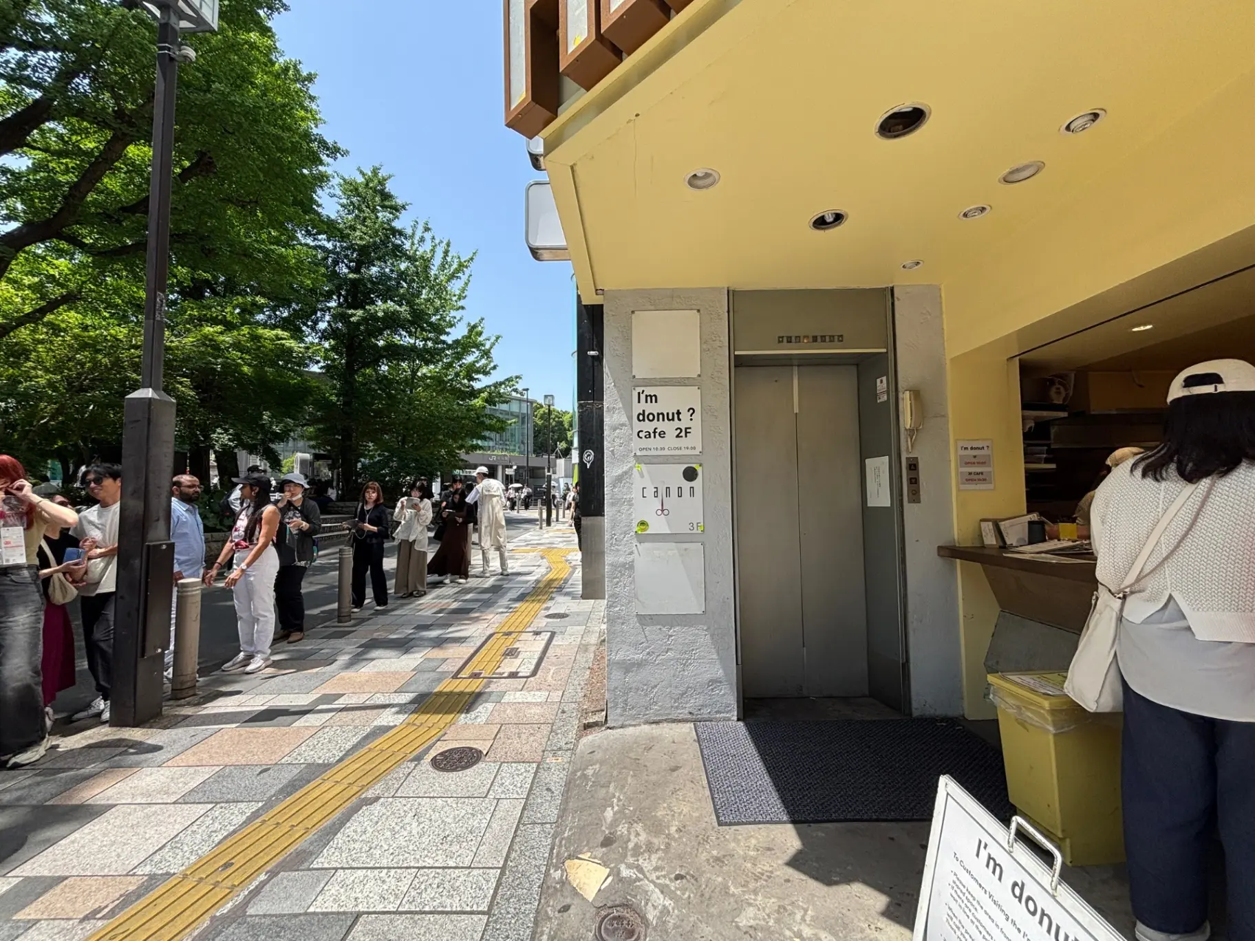 - 行列のあるカフェ入り口、明るい日差しの下で待つ人々と緑豊かな木々、階段を上るエレベーターの案内看板が目立つ街の風景