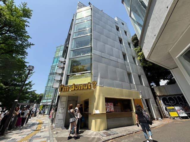 - モダンなデザインのカフェ「I'm donut?」、行列ができる人気の窓口、青空の下に立つメタリックな外観と緑の木々に囲まれたビル