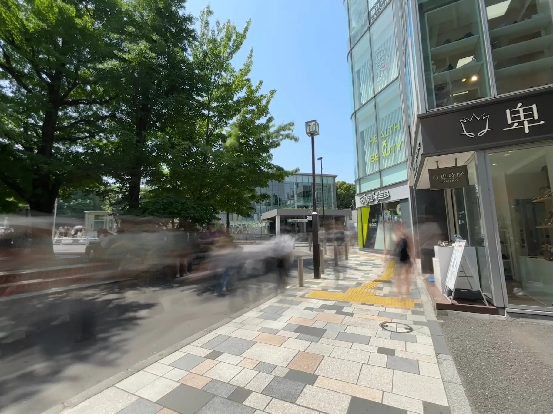 - 青空の下、繁忙な都市の通りに沿った歩行者と緑豊かな街路樹が見える風景、商業施設のガラス張りの外観とテラスが特徴的なショッピングエリア