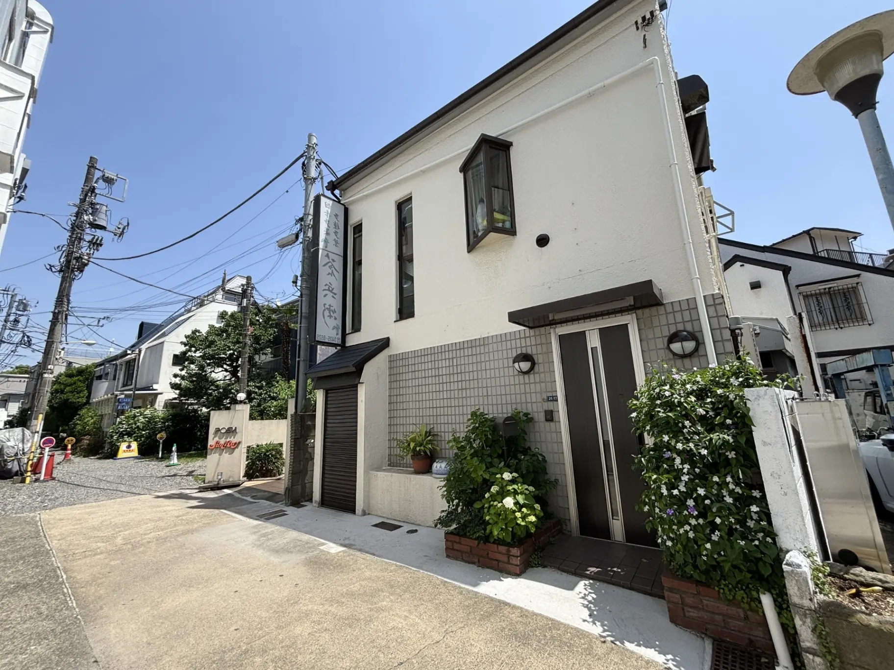 - 白い外壁の一戸建て住宅、屋外に植栽があり、周囲には電柱と電線が見える青空の下の風景