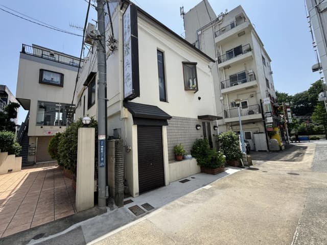 - 住宅街の一角にある白い外壁の建物、隣に高層ビルが立ち並ぶ様子、青空の下での風景