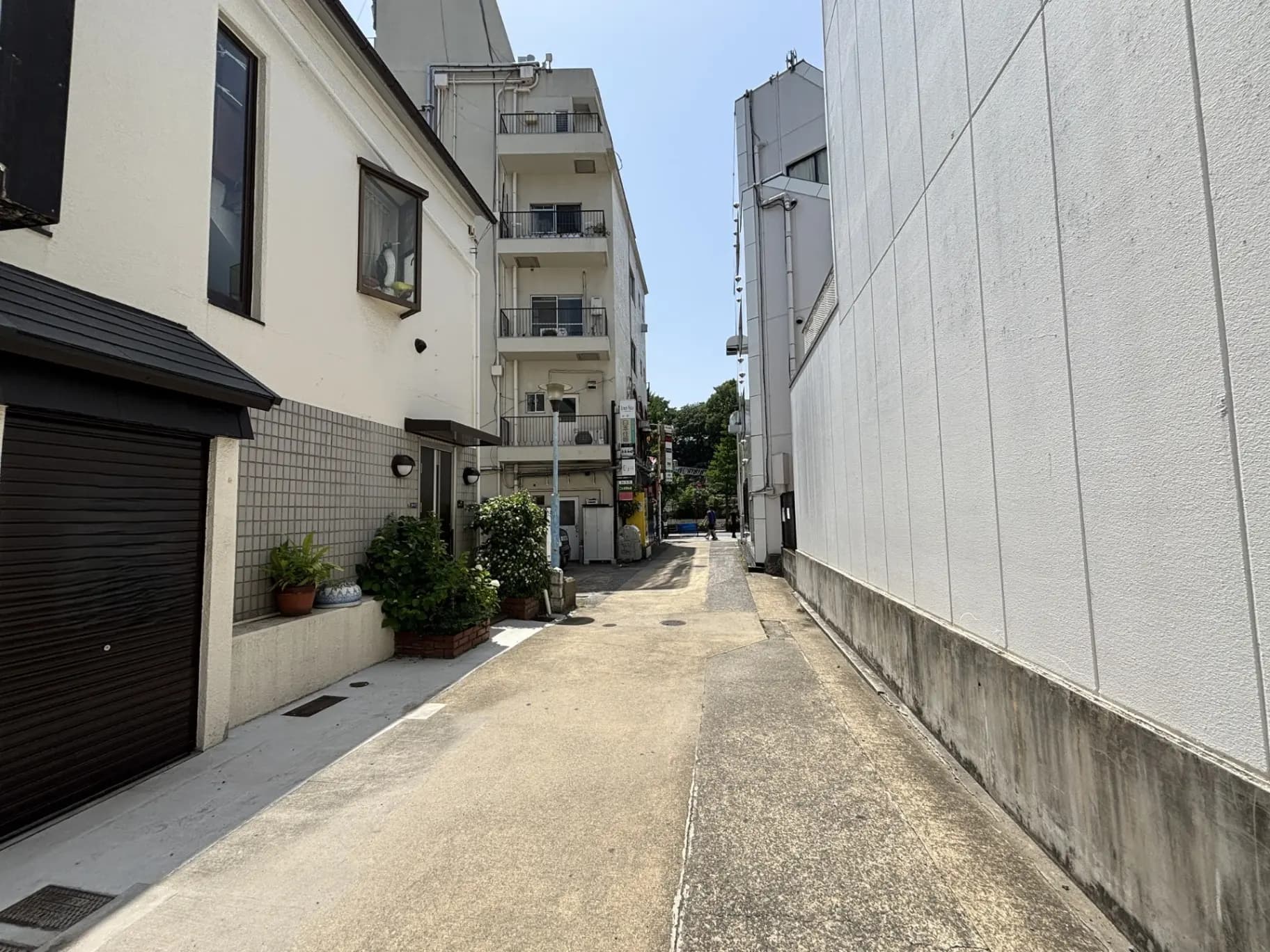 - 都市の住宅街に位置する狭い路地の風景、白い建物と黒いガレージ、植えられた草花が整然と並ぶ街並み